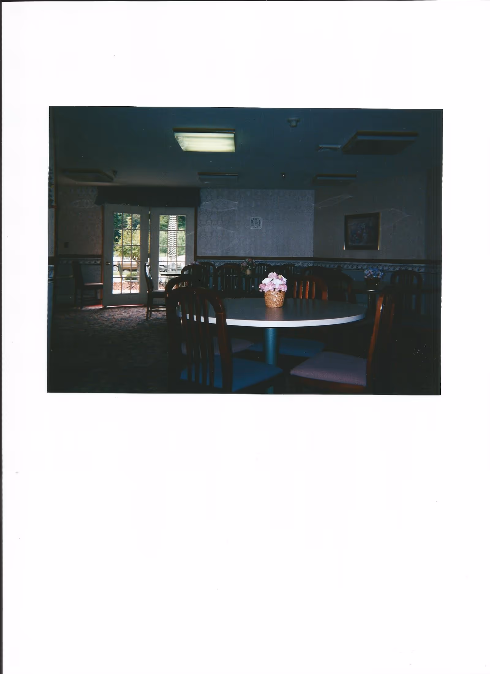 Interior view of a dining room with round tables and wooden chairs. Each table has a small basket with pink flowers. There are double glass doors at the back letting in natural light, and a framed picture on the wall.