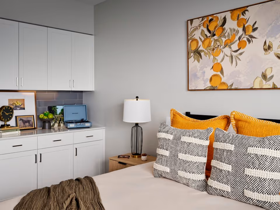 A cozy bedroom corner featuring a bed with decorative pillows in black, white, and mustard yellow. Next to the bed is a wooden nightstand with a modern lamp and a pair of glasses. On the left side, there is a white cabinet with drawers and cupboards, topped with framed pictures, a clock, a bowl of green apples, and a blue record player. Above the bed hangs a framed artwork depicting oranges and leaves.