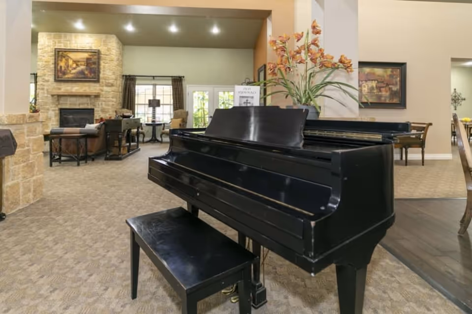 A black grand piano and bench sit in a spacious assisted-living common room with seating, a stone fireplace, and decorative flowers.