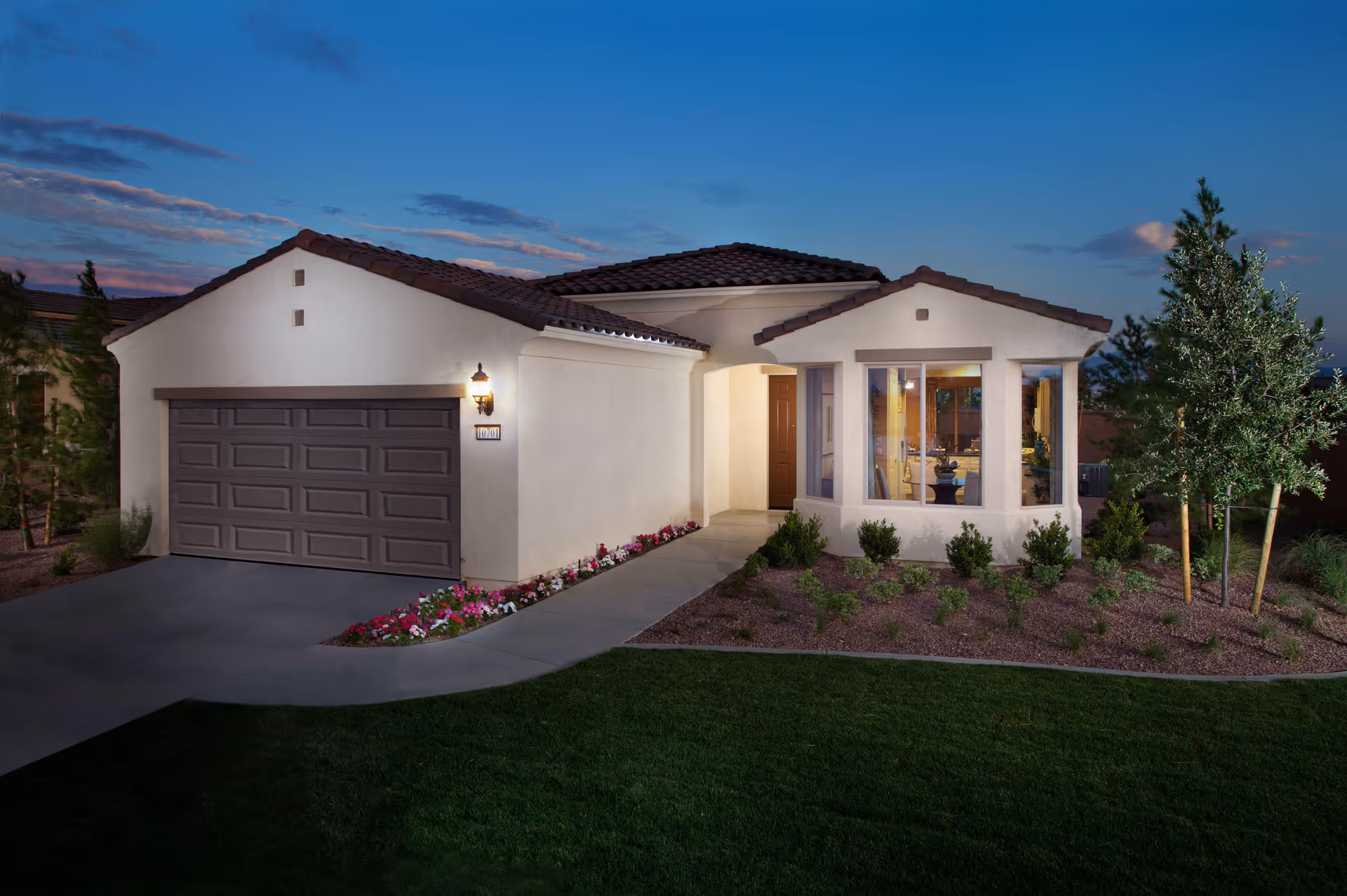 Exterior view of a single-story house at dusk with a two-car garage, a pathway leading to the front door, landscaped garden with small bushes and trees, and a well-maintained lawn.