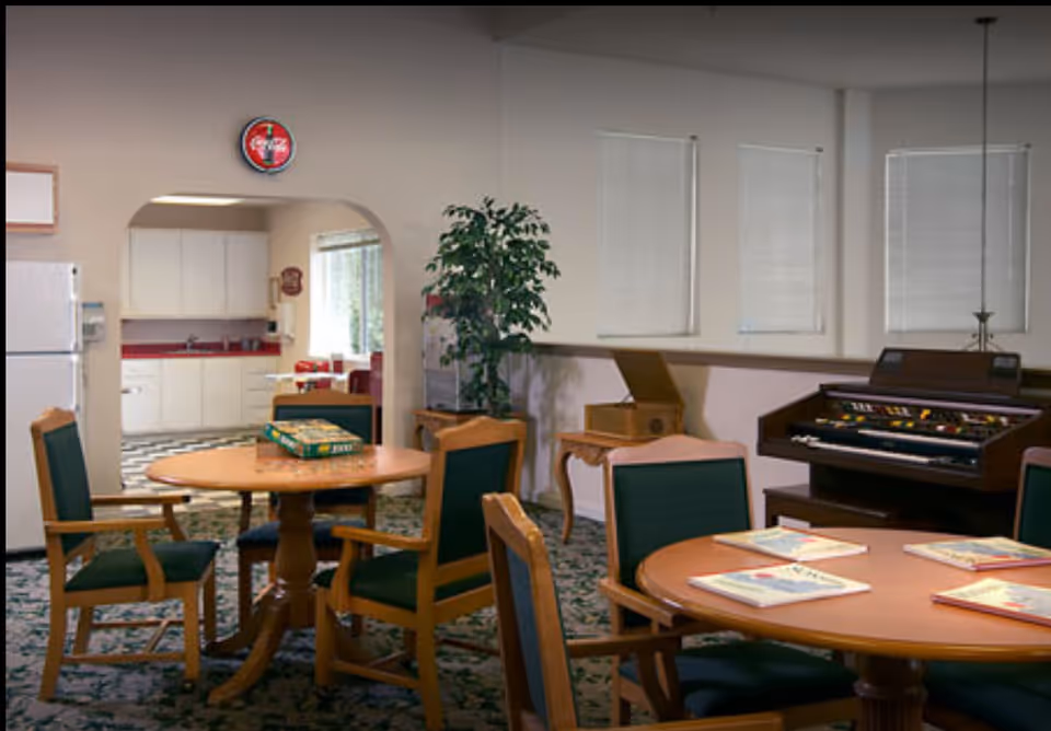 A common area in a senior living facility featuring two round wooden tables with green cushioned chairs. One table has a board game on it, and the other has several books. In the background, there is a kitchen area with white cabinets and a refrigerator. A vintage organ and a potted plant are also visible in the room.