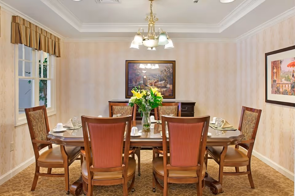 A dining room with a rectangular wooden table set for six people. The table has a glass top and is decorated with a vase of yellow flowers in the center. Six wooden chairs with cushioned seats and backs surround the table. The room has beige patterned wallpaper, a carpeted floor, two framed paintings on the walls, and a window with a striped valance. A chandelier with multiple lights hangs above the table.