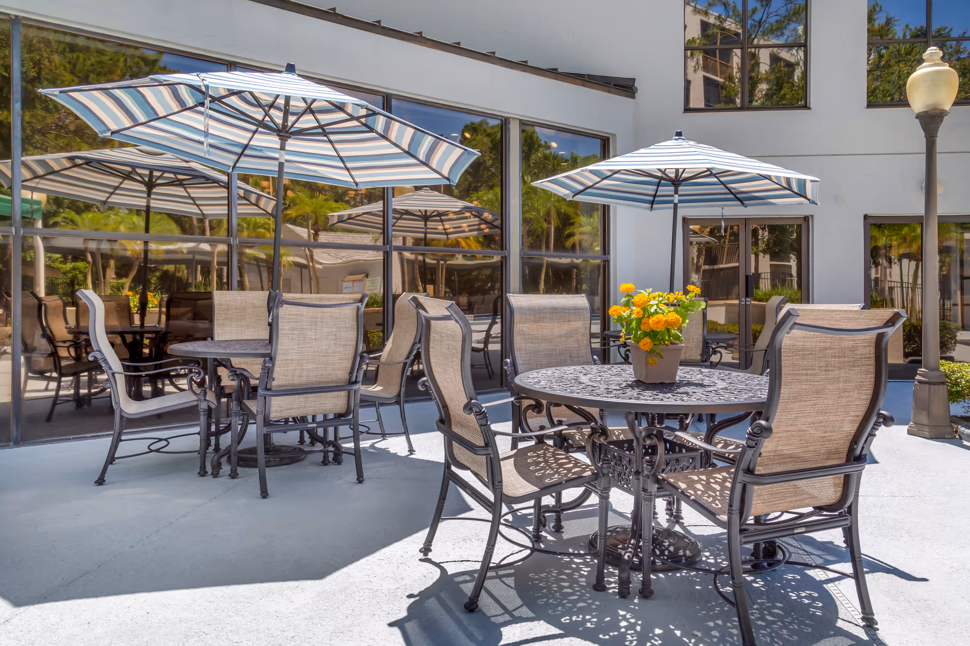 Outdoor patio area with round metal tables and cushioned chairs under striped umbrellas. A potted plant with yellow flowers is placed on one of the tables. The background shows large windows reflecting greenery and part of the building exterior.