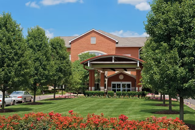Front exterior view of a red brick building with a covered entrance, surrounded by green trees, a well-maintained lawn, and red flowers under a blue sky with some clouds.