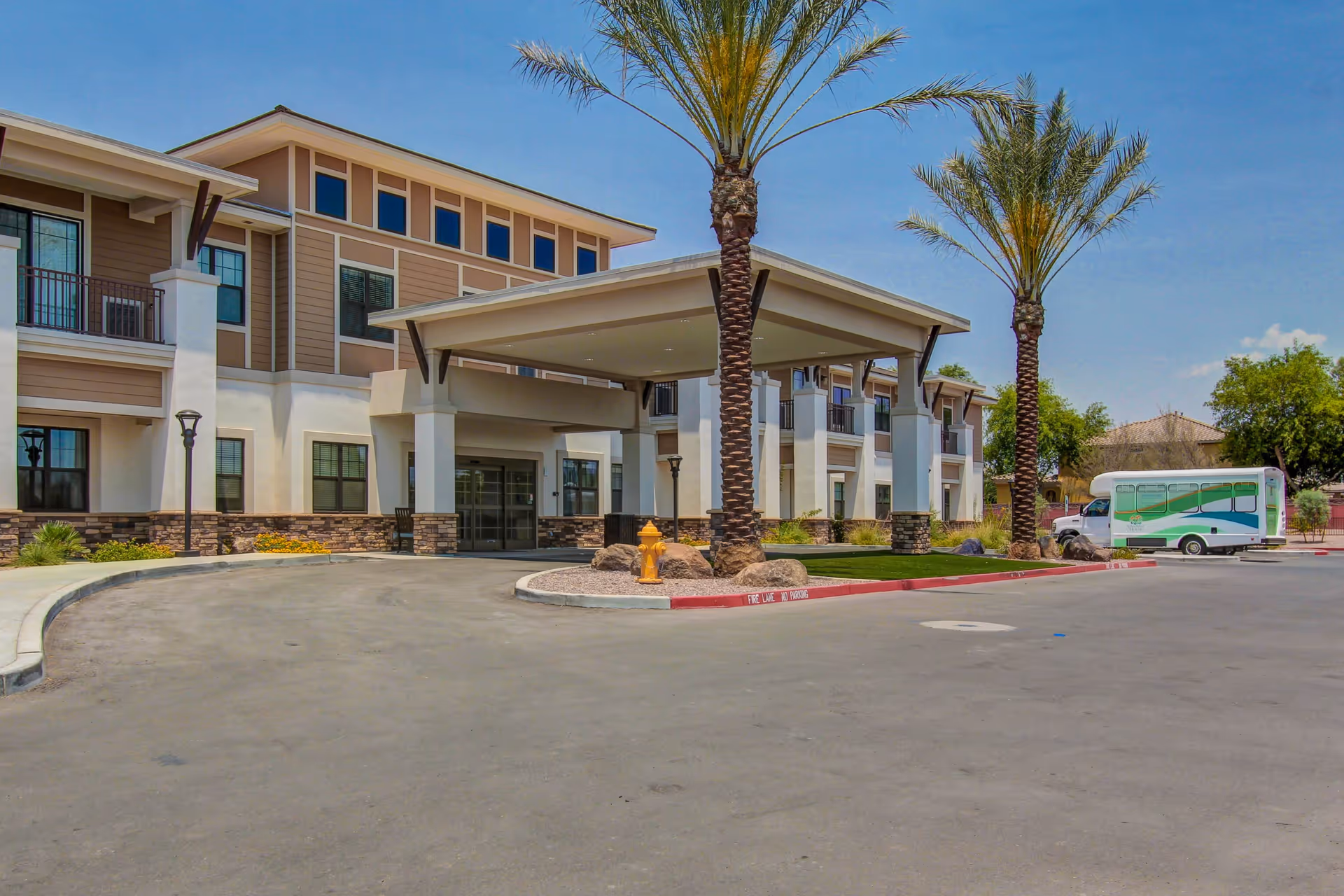 Exterior view of Savanna House senior living facility showing the main entrance with a covered drop-off area, palm trees, a fire hydrant, and a shuttle bus parked in the driveway under a clear blue sky.
