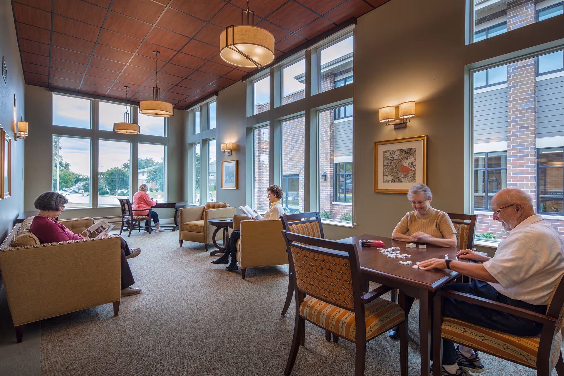A bright and spacious common area in a senior living facility with large windows letting in natural light. Several elderly people are seated comfortably; two are playing a game at a table, while others are reading or sitting on sofas and chairs. The room features warm lighting fixtures, framed artwork on the walls, and a carpeted floor.