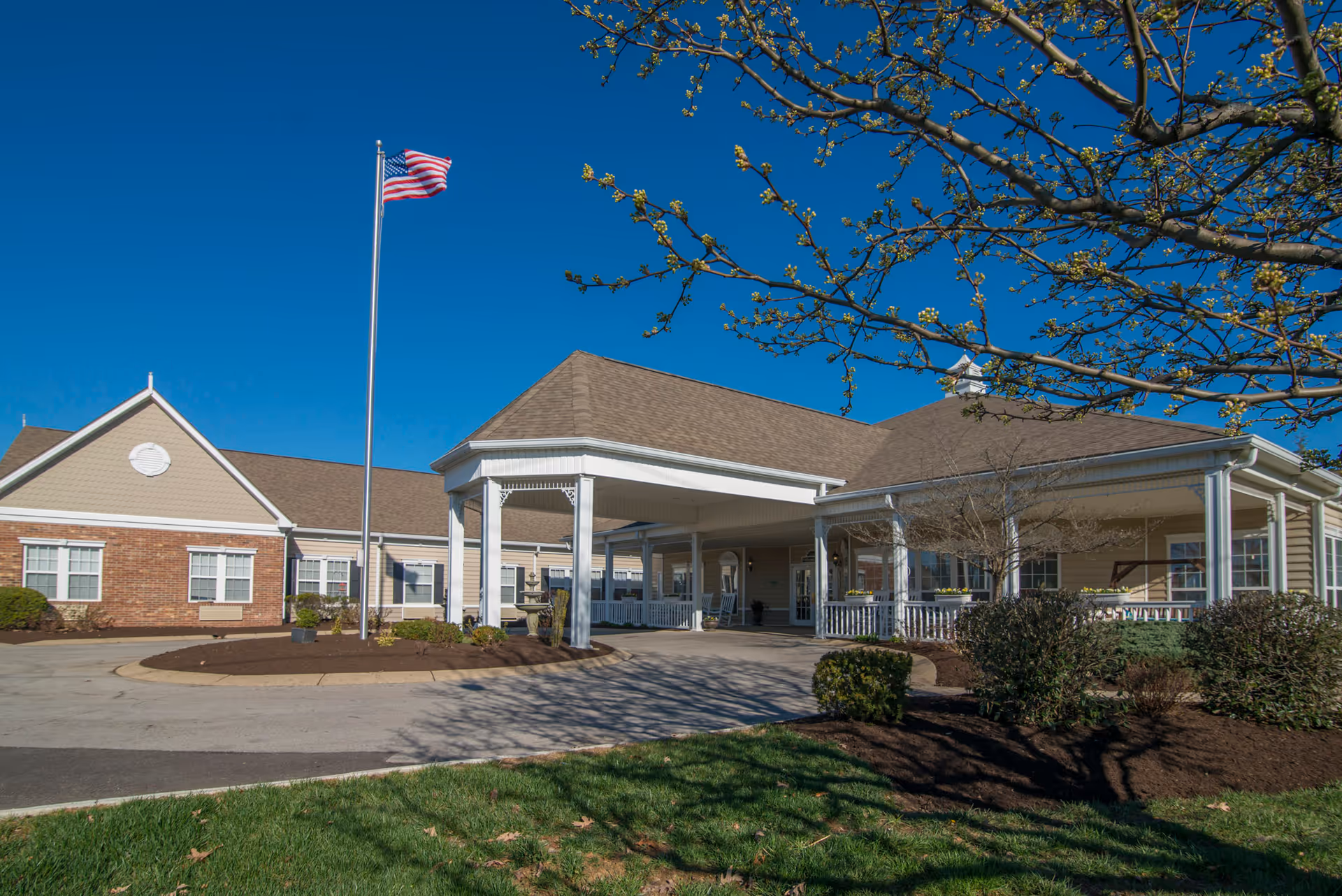 Exterior view of Morning Pointe of Frankfort building with a covered entrance, an American flag on a flagpole, and landscaping including bushes and a tree with budding leaves under a clear blue sky.