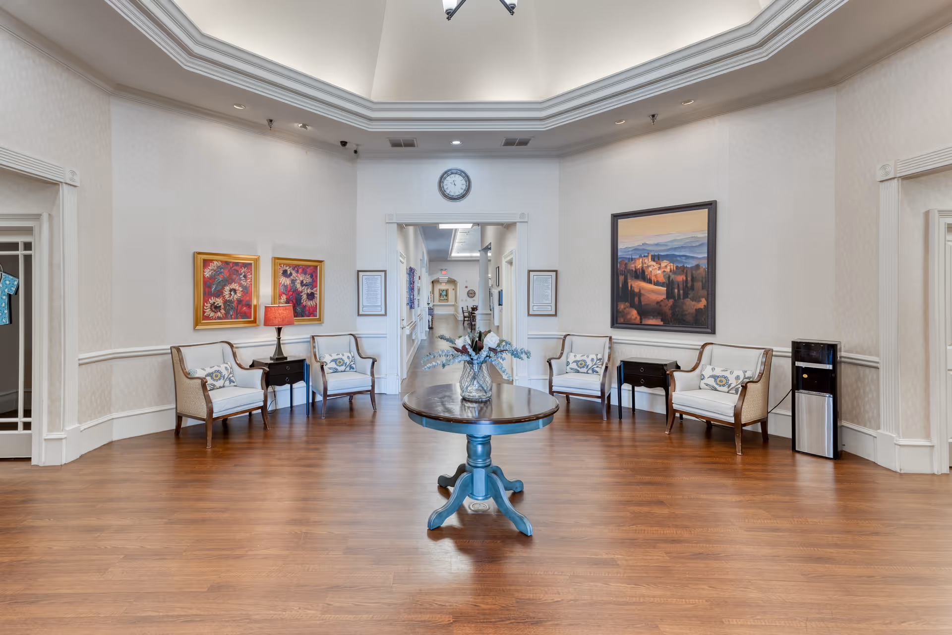 A spacious, well-lit common area with wooden flooring and light-colored walls. The room features a round wooden table with a floral arrangement in the center. Along the walls are several cushioned armchairs with decorative pillows, small side tables, framed artwork, and a clock above a doorway leading to a hallway. A water cooler is positioned on the right side.