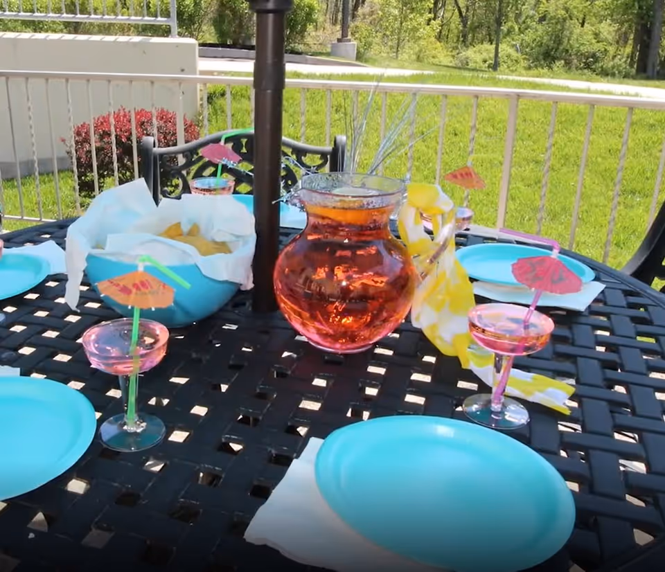 Outdoor patio table set with blue plates, a bowl of chips, a pitcher of pink beverage, and pink cocktail glasses with small paper umbrellas and green straws. The table is black metal with a lattice design and is surrounded by a white railing with green grass and trees in the background.