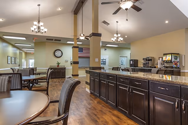 Interior view of a senior living facility dining area with round tables and chairs, a long granite countertop with dark wood cabinets, ceiling fans, chandeliers, and a beverage station with coffee and juice dispensers. An American flag and a clock are visible on the far wall.
