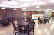 A spacious interior common area with multiple round tables and chairs arranged throughout the room. The walls are light-colored with a brick accent wall on one side. The ceiling has several round light fixtures, and there is a reception or service desk visible in the background.