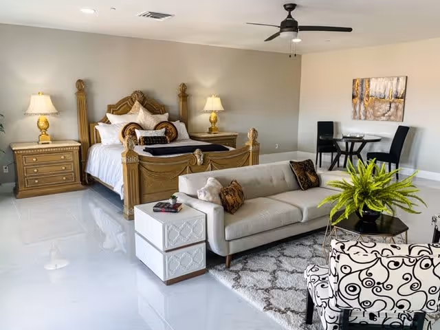 A bright studio-style suite featuring an ornate wooden bed flanked by lamps, a sofa and armchair, a small dining table, and a ceiling fan on a glossy tile floor.