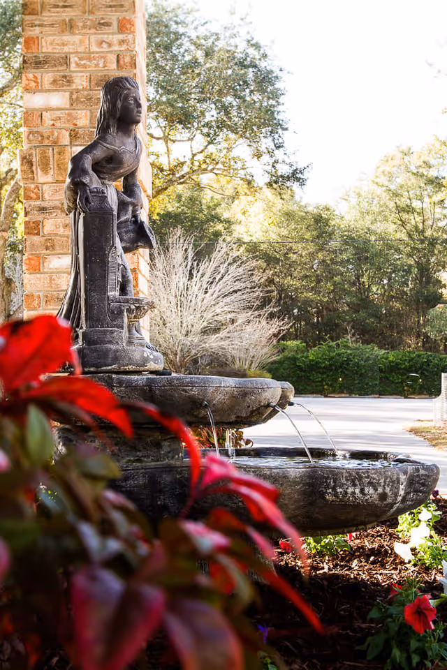 Outdoor stone water fountain with a statue of a girl holding a basket, surrounded by red and green plants, with trees and a driveway in the background.