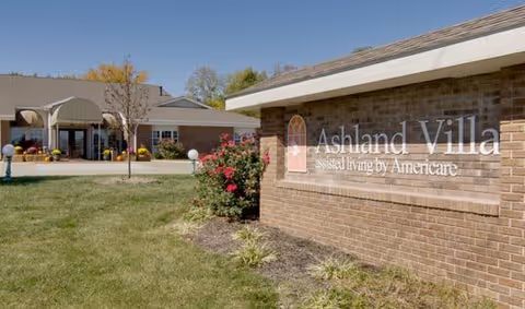 Exterior view of Ashland Villa assisted living facility showing a brick sign with the facility name and a building entrance in the background surrounded by grass and some landscaping.