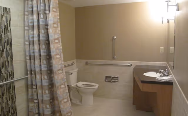 A bathroom with beige tiled walls and floor, featuring a toilet with grab bars on the walls around it, a shower area with a patterned shower curtain, and a sink with a countertop and mirror above it. The lighting is warm and there are two wall-mounted light fixtures above the sink.
