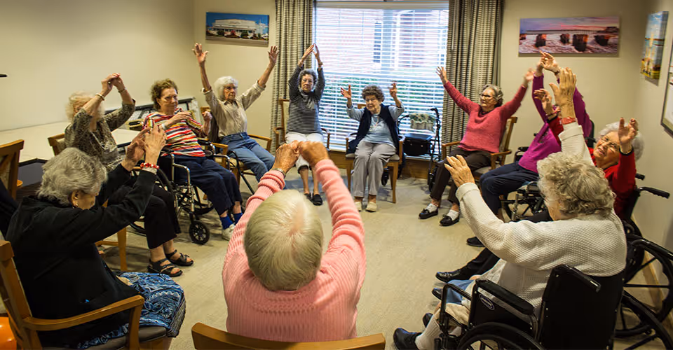 A group of elderly women sitting in a circle in a room, participating in a seated exercise or stretching activity with their arms raised. Some are in wheelchairs, and the room has a large window with blinds and curtains, along with framed pictures on the walls.