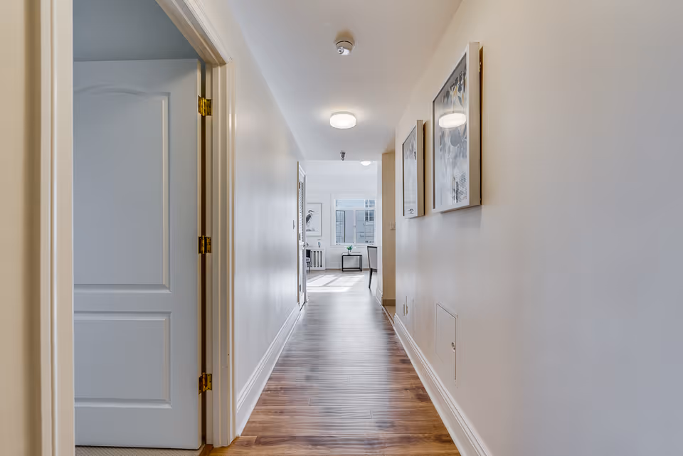 A long hallway with wooden flooring and white walls. On the left side, there is a partially open white door. On the right wall, there are two framed pictures. At the end of the hallway, a bright room with large windows and a small table with a flower vase is visible.