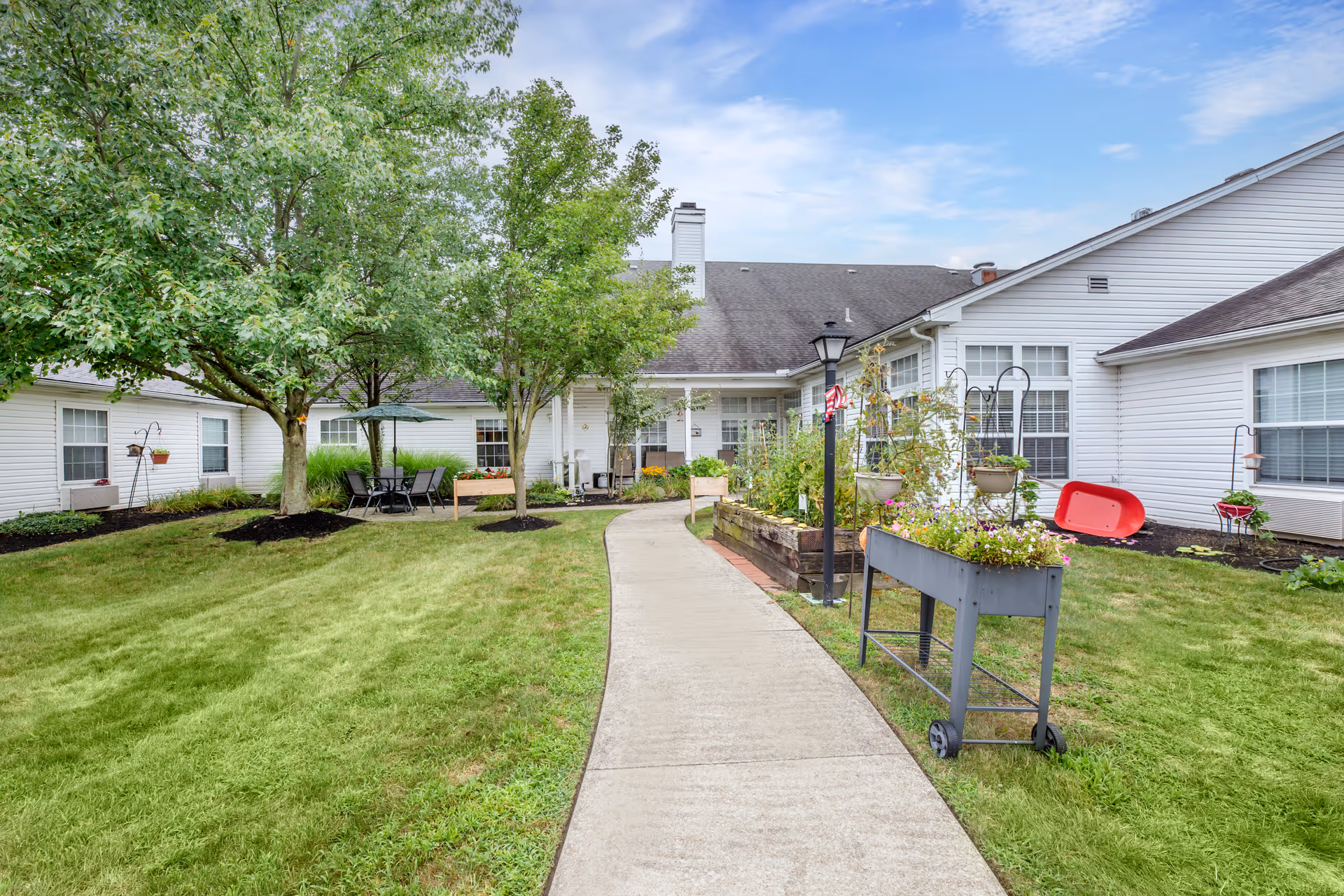 A well-maintained outdoor garden area at Brookdale Barberton featuring a concrete walkway, green grass, trees, raised garden beds with flowers and plants, a patio table with chairs and an umbrella, and white residential-style buildings surrounding the garden.