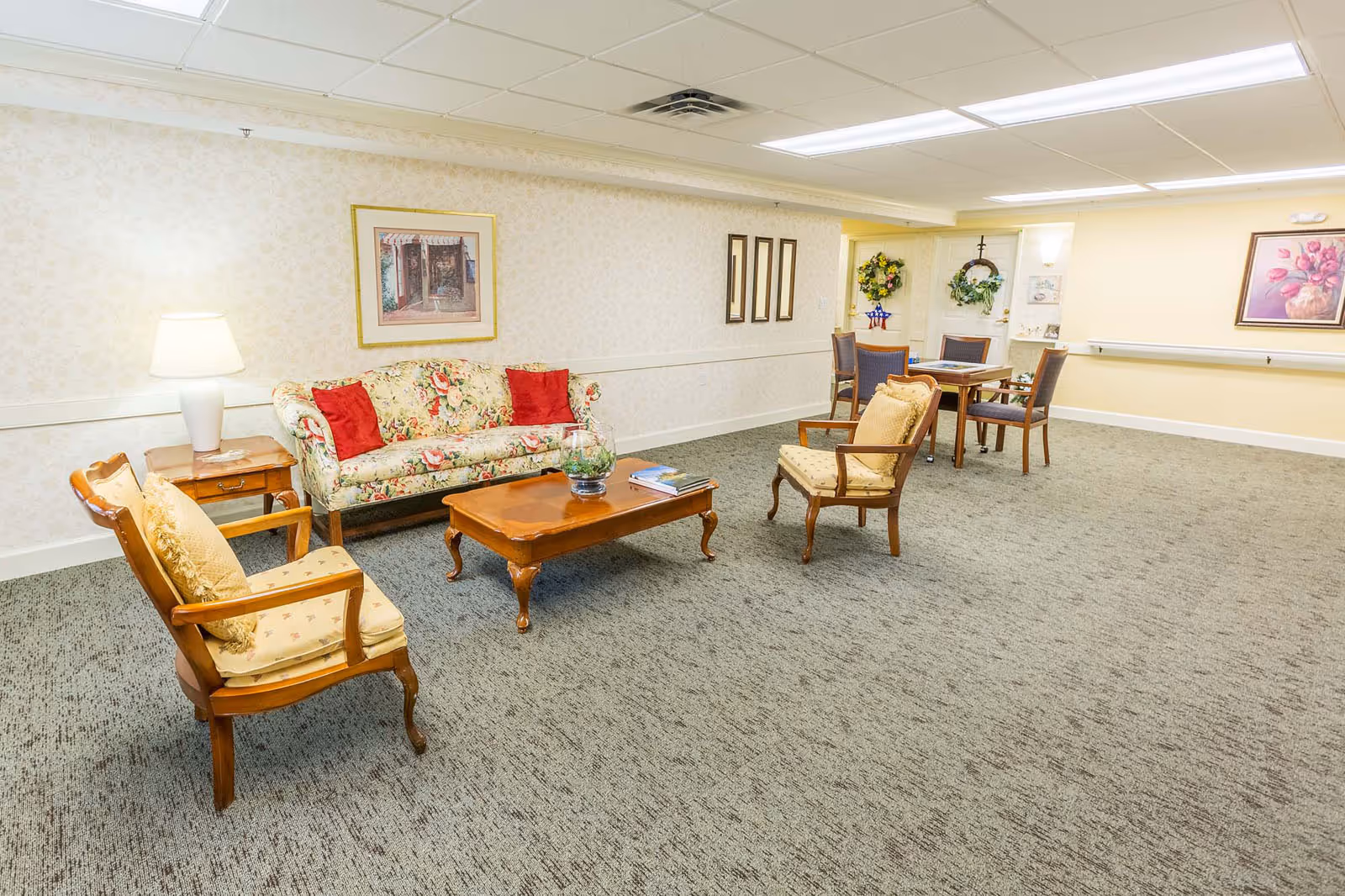 A spacious senior living common room with a floral sofa, armchairs, a coffee table and a small dining table.