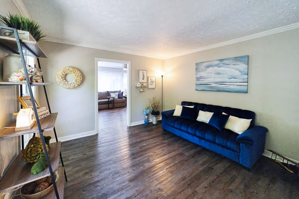 A cozy living room with a dark blue sofa adorned with light-colored pillows, a floor lamp in the corner, and a large painting of a cloudy sky above the sofa. To the left, there is a tall shelving unit with various decorative items and plants. The room has wood flooring and light gray walls. An open doorway leads to another room with a brown sofa and sheer curtains.