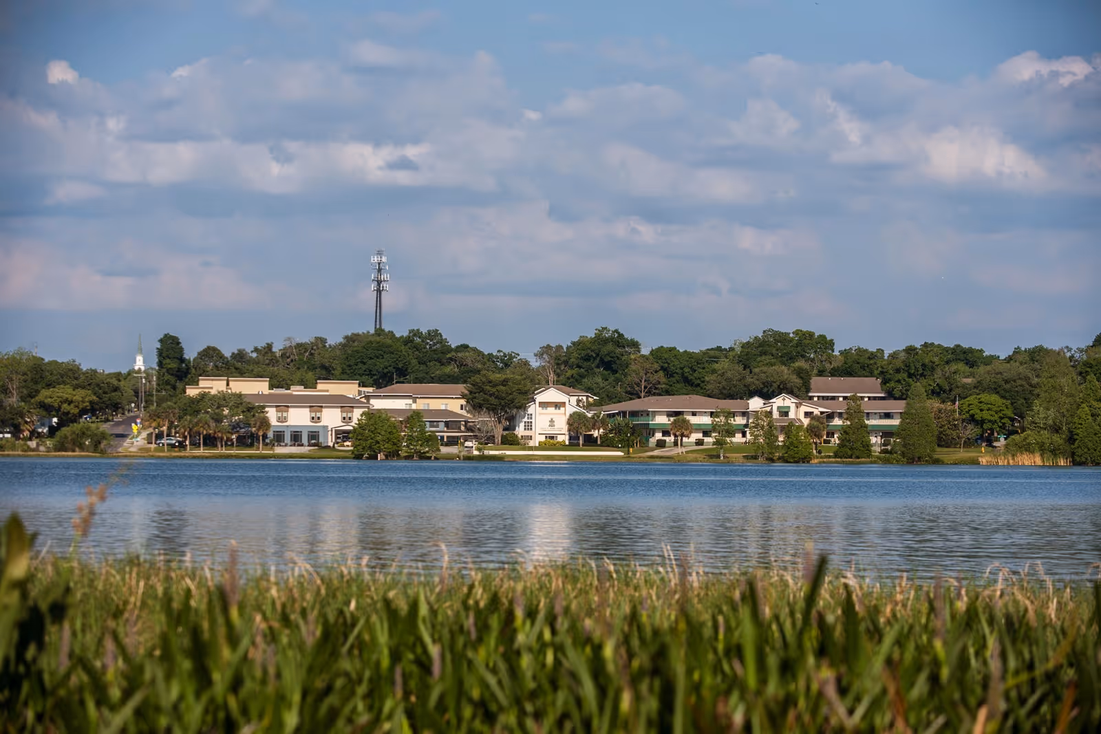 Buildings of a senior living campus seen across a lake with grassy foreground, trees, and a cloudy sky.