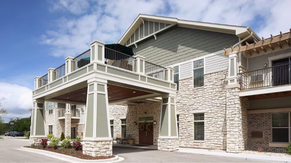 Entrance of a two-story assisted living building with a covered porte-cochere, stone facade, balconies and landscaping under a partly cloudy sky.