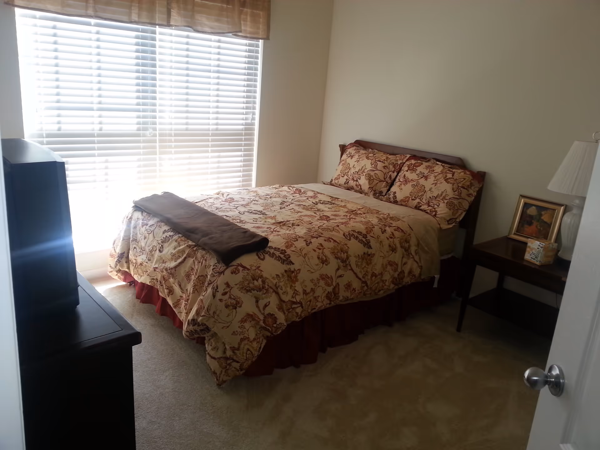 A bedroom with a bed covered in a floral patterned comforter and matching pillows. There is a brown blanket folded at the foot of the bed. A window with white blinds and a valance is behind the bed. To the right of the bed is a wooden nightstand with a white lamp, a framed picture, and a box of tissues. A dark-colored dresser with a television on top is positioned to the left of the bed. The room has beige carpet and light-colored walls.