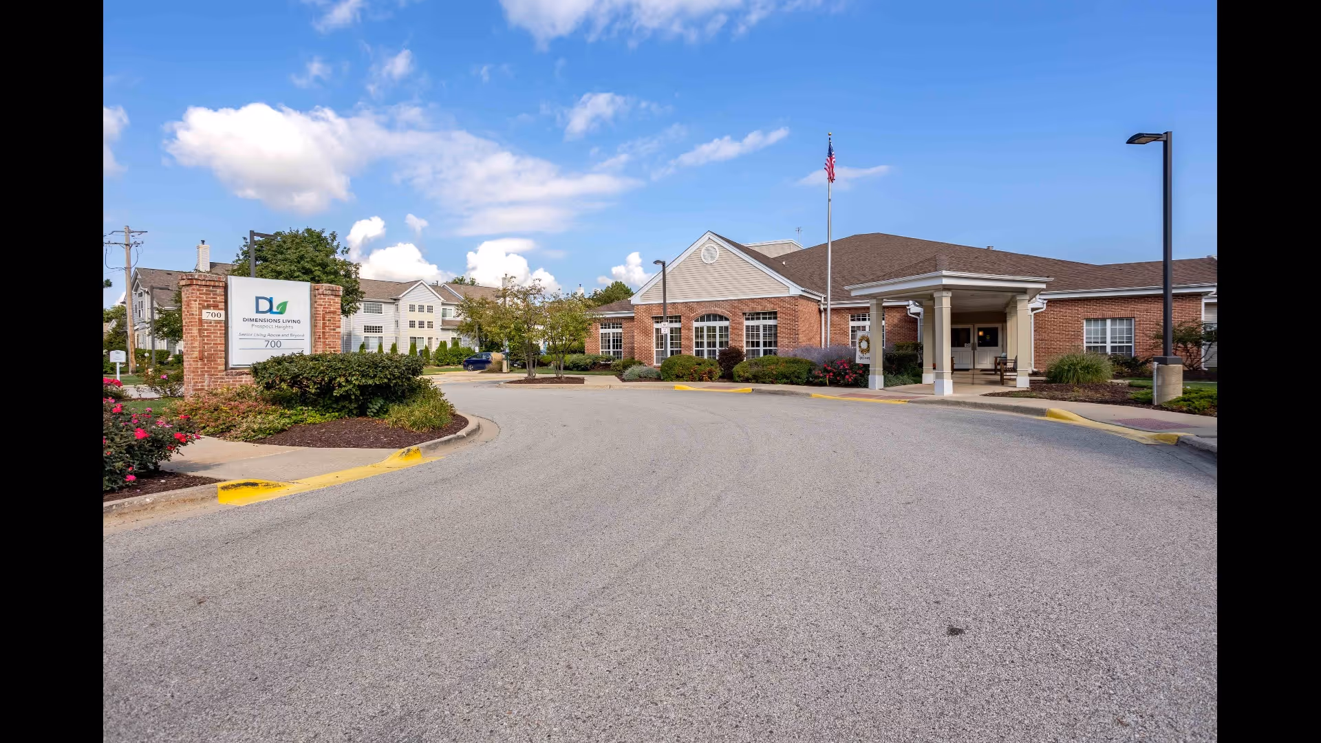 Front entrance of a one-story brick senior living building with a circular driveway, flagpole, landscaping, and a sign near the curb.
