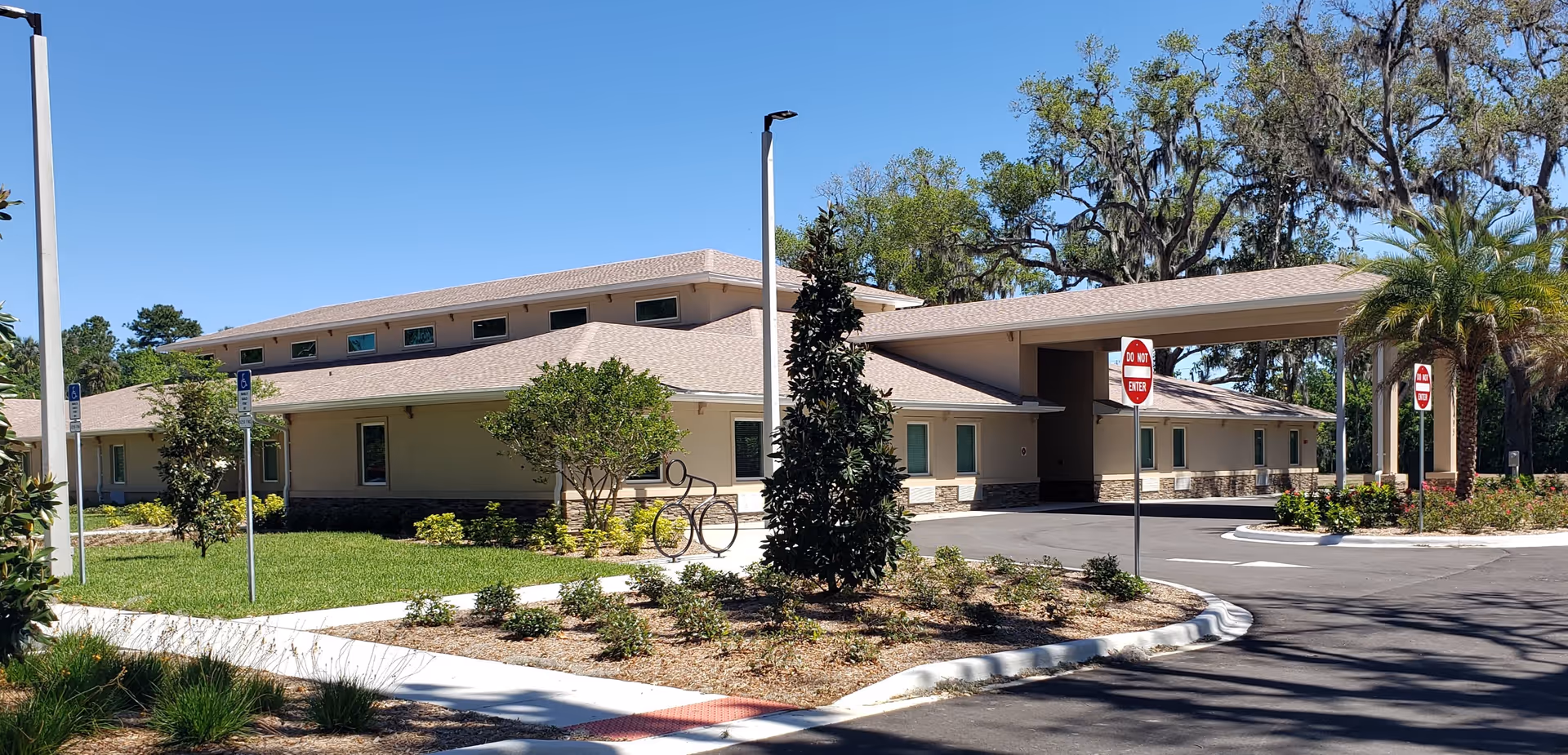 Exterior view of a single-story building with beige walls and a tan roof under a clear blue sky. The building has multiple windows and a covered entrance with a driveway. There are landscaped areas with green grass, small bushes, trees, and a palm tree. Several 'Do Not Enter' signs and handicap parking signs are visible along the driveway.