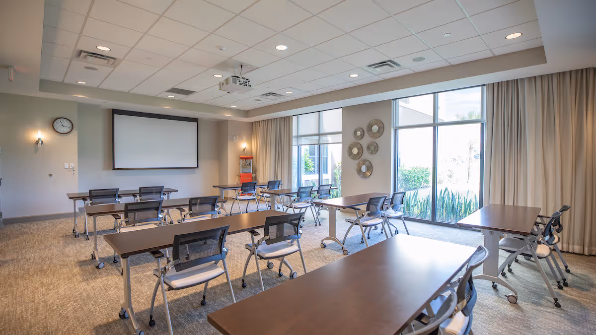 A bright and spacious meeting or classroom with multiple tables and chairs on wheels arranged in rows facing a large projection screen on the wall. The room has large windows with beige curtains allowing natural light to enter, a wall clock, and decorative round mirrors on one wall. There is also a popcorn machine in the corner.