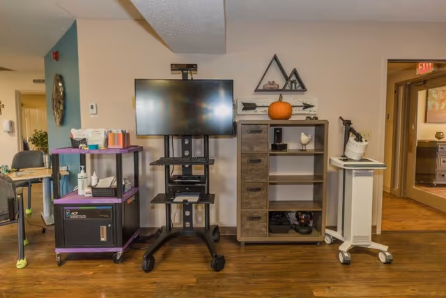 An interior room with a mounted flat-screen TV on a black stand, a purple and black utility cart with various supplies, a wooden cabinet with shelves and drawers decorated with a pumpkin and small ornaments, and a white medical equipment cart on wheels. The room has wood flooring and beige walls, with a hallway and door visible in the background.