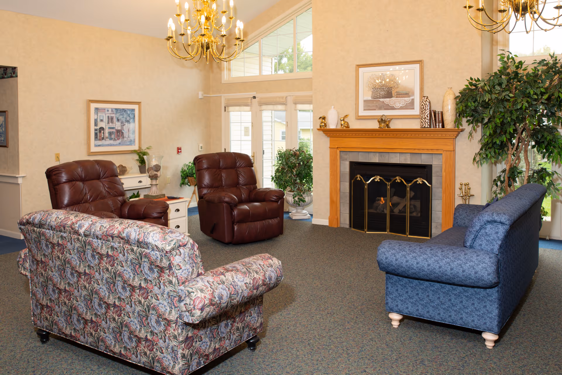 A cozy living room with two patterned sofas, two brown leather recliners, a wooden fireplace with a fire burning, decorative items on the mantel, a large plant, framed artwork on the walls, and a chandelier hanging from the ceiling. There are large windows and glass doors letting in natural light.