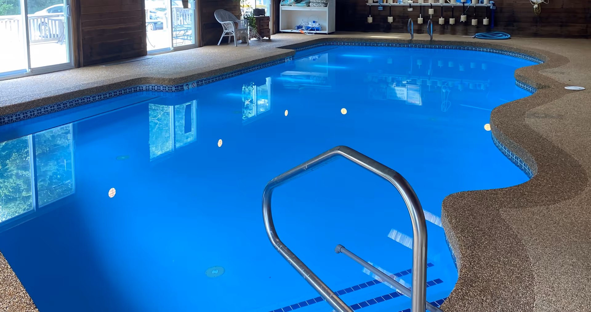 Indoor swimming pool with clear blue water, metal handrail, and textured pool deck. Large windows and sliding glass doors allow natural light to enter the room. Chairs and pool equipment are visible in the background.