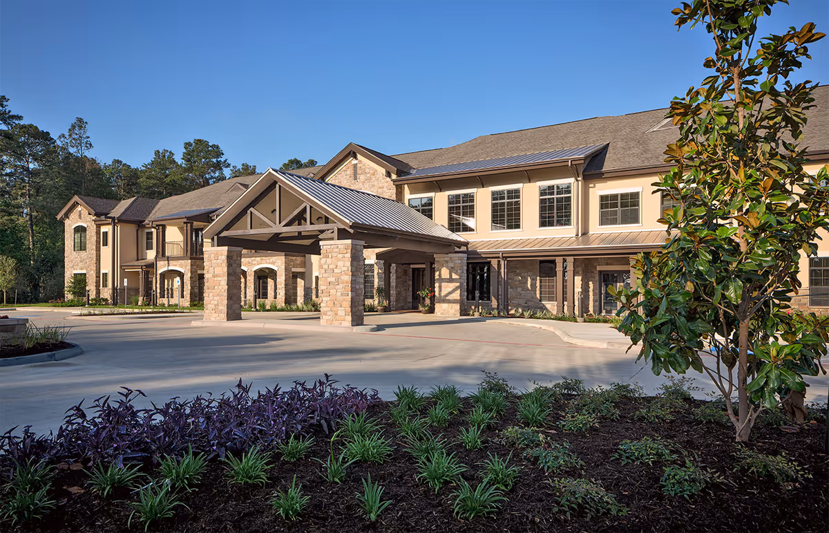 Exterior view of a senior living facility named Spring Creek Village, featuring a two-story building with stone and beige siding, a covered entrance supported by stone pillars, landscaped greenery, and a clear blue sky.