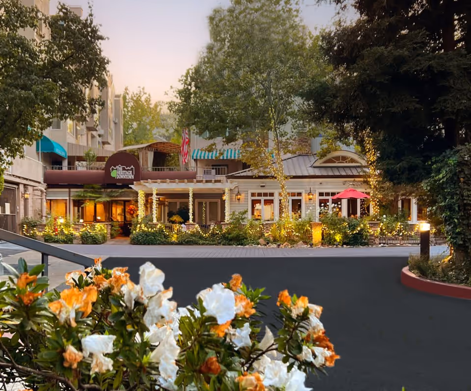 Exterior view of The Heritage Downtown senior living facility at dusk, featuring a well-lit entrance with decorative string lights wrapped around trees and columns. The building has a welcoming porch area with greenery and flowers in the foreground, and an American flag is visible near the entrance.
