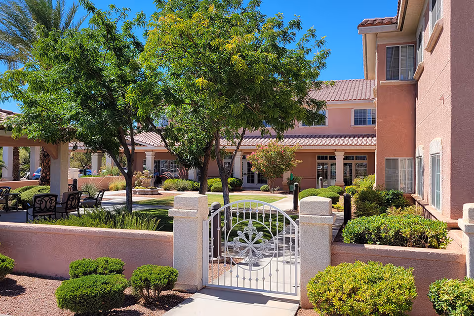 Outdoor courtyard area of a senior living facility with a gated entrance, green trees, shrubs, patio seating with chairs, and a pink stucco building with tiled roof in the background under a clear blue sky.
