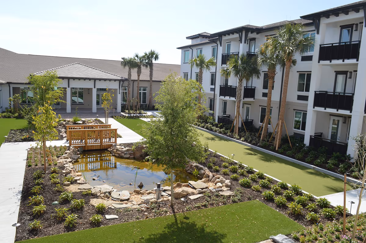 Landscaped courtyard with a small pond and wooden footbridge, surrounded by palm trees and a three-story senior living building.