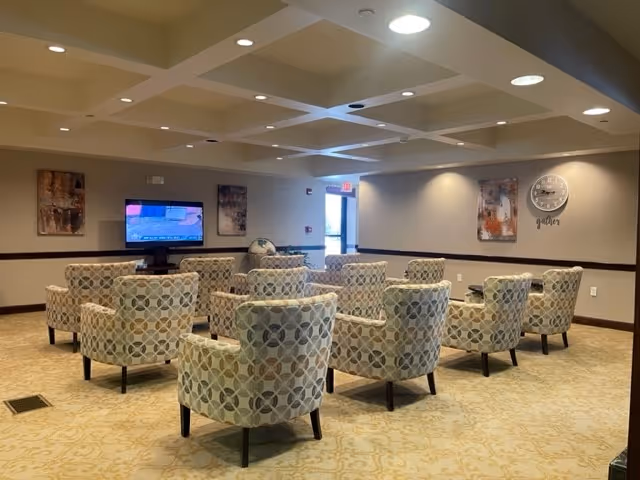 A senior living facility common room with rows of patterned armchairs arranged facing a wall-mounted television.