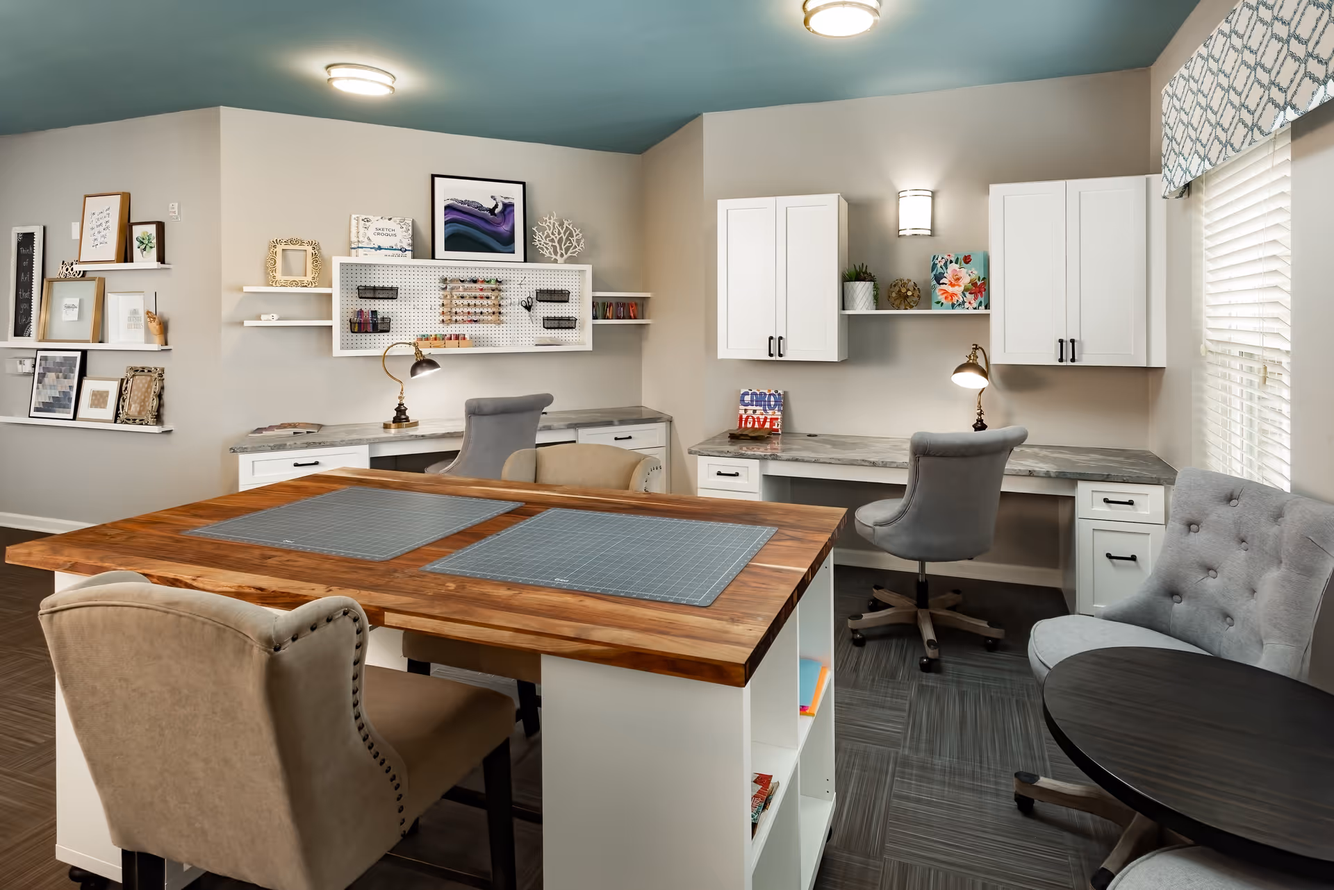 Bright interior activity room with a central wooden work table, upholstered chairs, built-in desks, cabinets, and shelving.