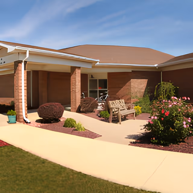 Front entrance of a single-story brick senior living building with a covered walkway, bench, landscaping and flowering bushes.
