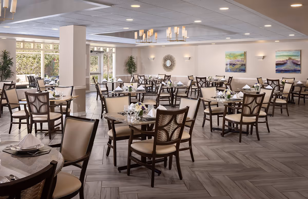 Spacious dining room with multiple tables and chairs neatly arranged. Each table is set with white napkins, glassware, and small floral centerpieces. The room features large windows allowing natural light, modern ceiling lights, decorative wall art, and plants in the corners.