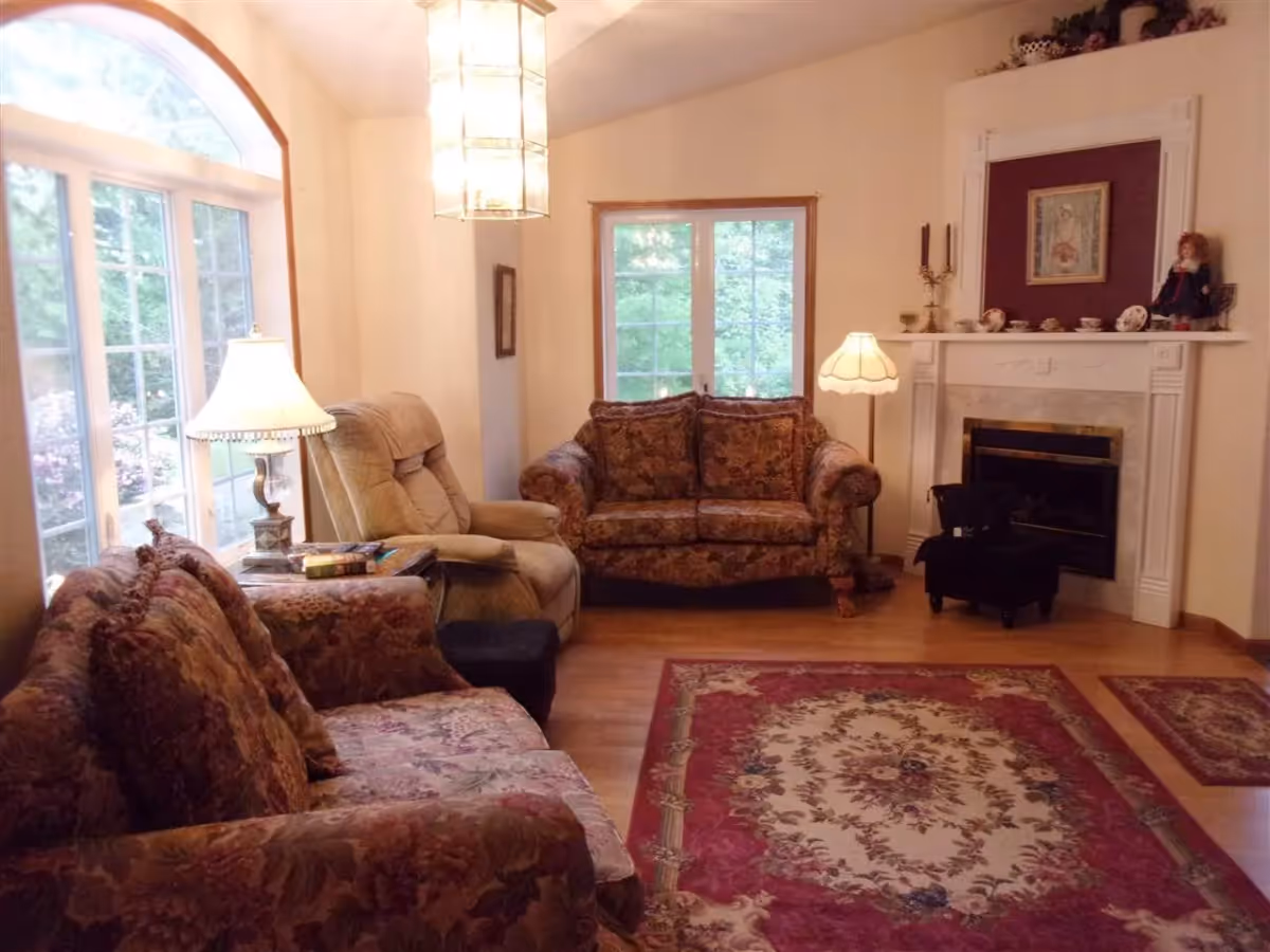 A cozy living room with floral patterned sofas, a beige recliner chair, a large window with an arched top letting in natural light, a decorative fireplace with various ornaments and a doll on the mantel, two standing lamps, and a red and beige patterned area rug on a wooden floor.