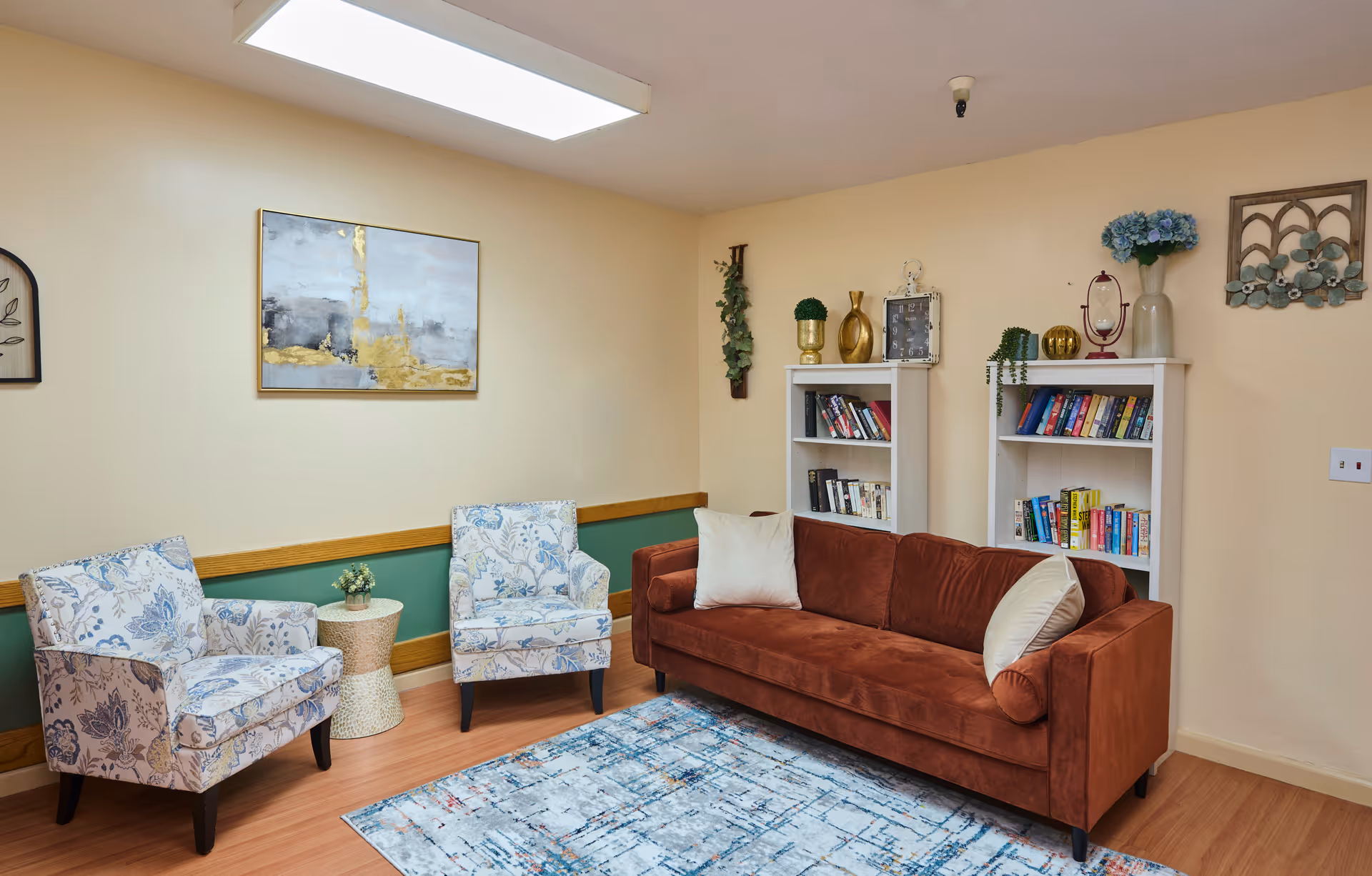 Cozy common room with a rust-colored sofa, two patterned armchairs, bookshelves filled with books, and wall decor.