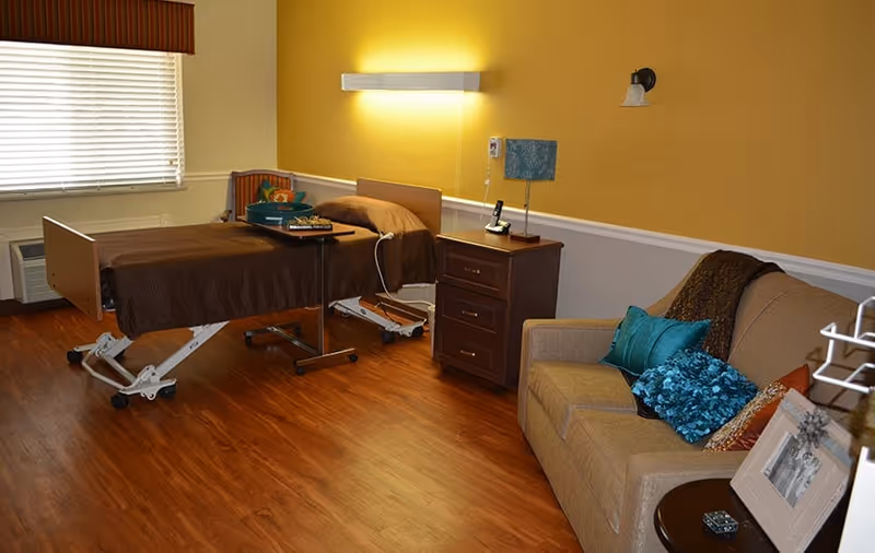 A senior living bedroom with an adjustable hospital-style bed, nightstand, sofa with decorative pillows, and warm yellow walls.