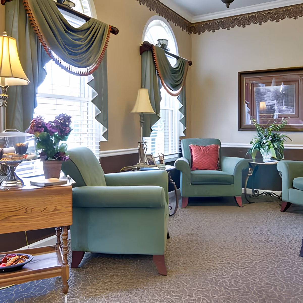 A cozy living room with green upholstered armchairs, a wooden side table with a potted plant and snacks, two tall windows with decorative green curtains, two standing lamps, a framed picture on the wall, and a carpeted floor.