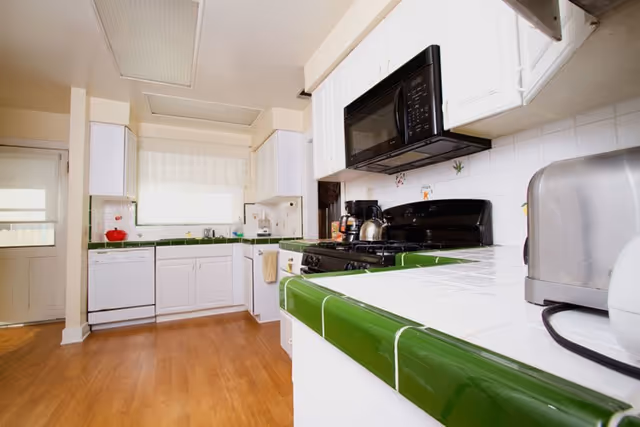 Bright kitchen with white cabinets and green tiled countertops. The kitchen features a black microwave above a black stove, a coffee maker, a toaster, and a dishwasher. There is a window with a sheer curtain above the sink, and the floor is wooden. The kitchen has a door with a window on the left side.