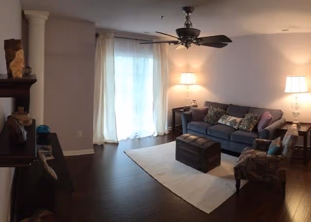 Cozy living room with a sofa, patterned armchair, trunk-style coffee table on a rug, ceiling fan, lamps, and sheer curtains over sliding doors.