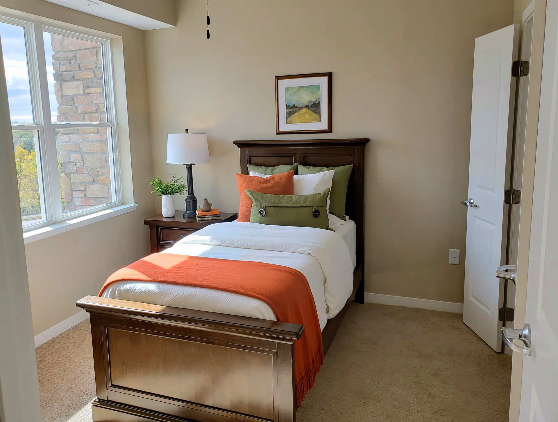 Sunlit single bedroom with a wooden bed dressed in white linens and an orange throw, a nightstand with a lamp, and a window showing a stone exterior.