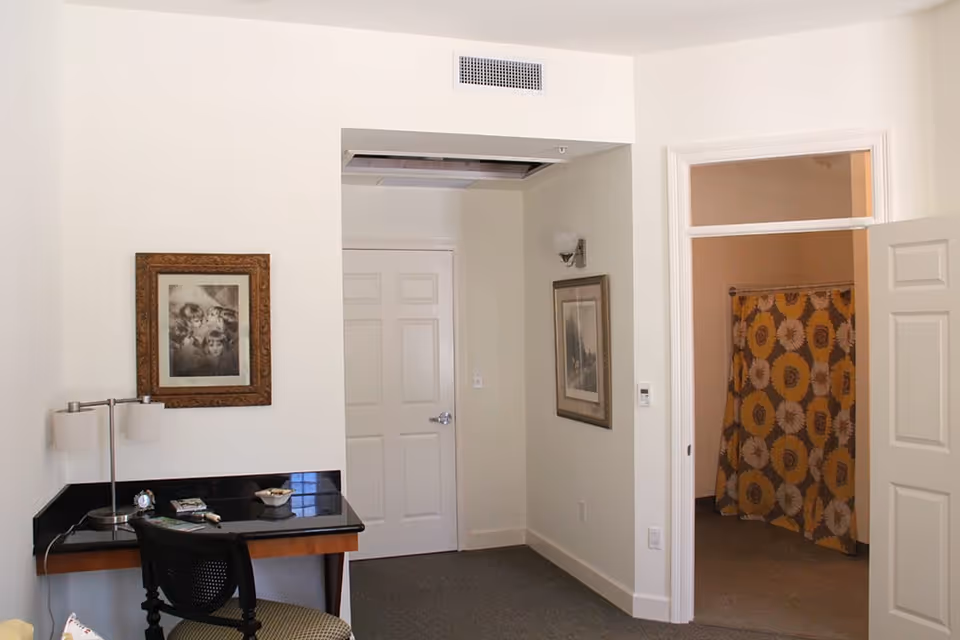 Interior view of a room with a small desk and chair on the left, a framed picture on the wall above the desk, a closed white door in the center, and an open doorway on the right revealing a bathroom with a yellow and white floral shower curtain.