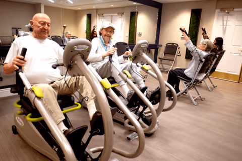 Four elderly individuals exercising on stationary recumbent bikes and resistance machines in a well-lit fitness room with wooden flooring and beige walls.